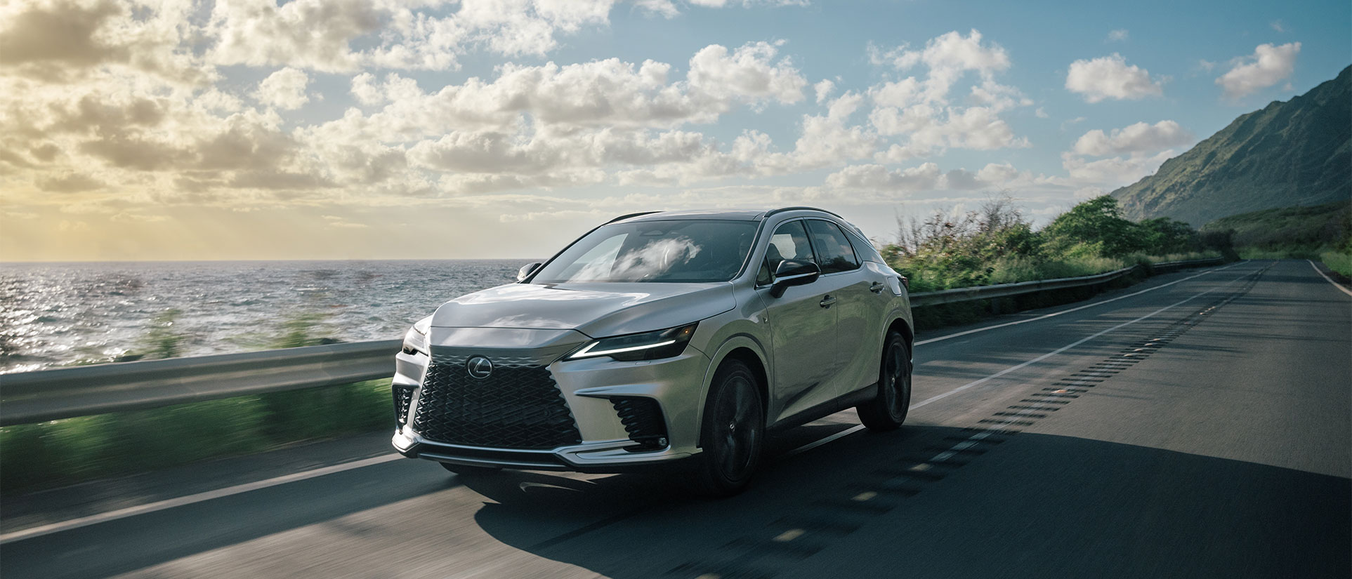 Silver Lexus RX with F Sport accentuated features driving down ocean-side highway under bright skies